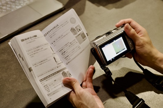 カメラのダイヤルを手で操作し、横にマニュアル本やスマホを置く学習風景